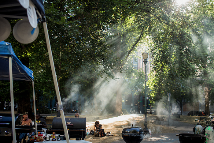 Smoke from four grills waft into Lownsdale Square with afternoon sun spilling through the trees to create rays of light and smoke.