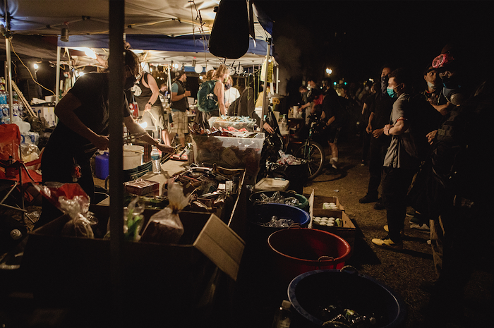 A volunteer organizes individually packaged snacks. Below the table, plastic bins are filled with ice and beverages. Protestors look on, debating what to snack on.