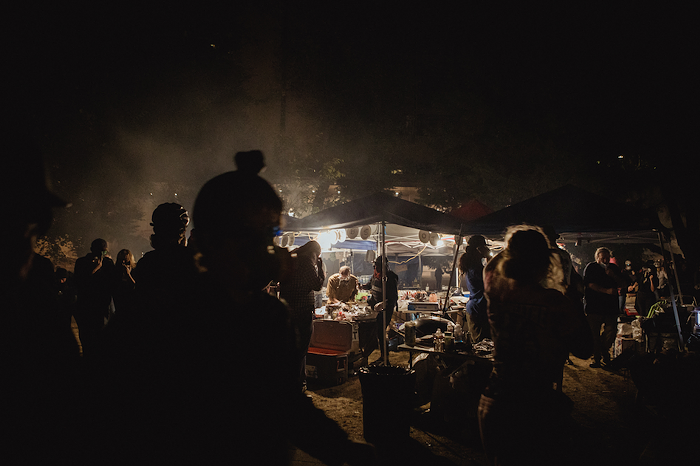 Riot Ribs volunteers continue to serve plates of food under a lit canopy. In the foreground, protestors in gas masks are silhouetted as they walk past to escape tear gas.