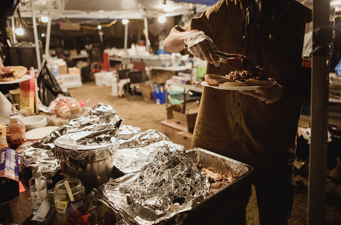 A volunteer wearing a gas mask and gloves holds a plate with buns with one hand and dishes barbecue with the other. Foil-covered serving trays full of food stand by, ready to be served.