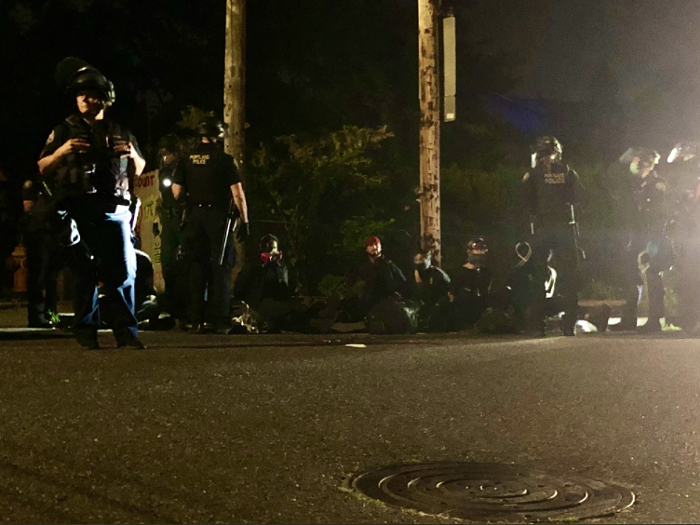 Seven arrestees are seated and handcuffed on a curb. (After this photo was taken, officers pushed all press out of the area.)