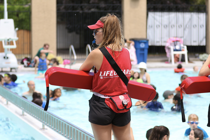 A lifeguard looks over the pool. They have a whistle in their mouth and are holding a flotation device.