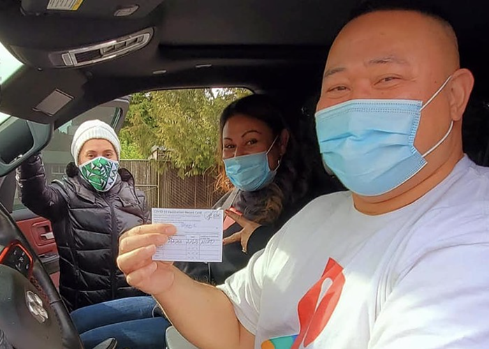 Two Pacific Islander people in a car wearing masks. One of them holds up their vaccination card.