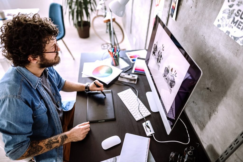 A man with curly hair and tattoos works on an illustration on his desktop computer. 