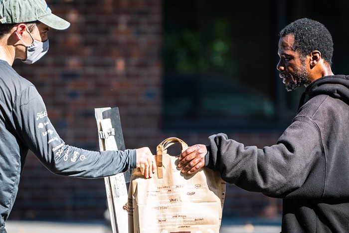A man with a mask hands a paper sack to another man, who is obviously sweating in the heat